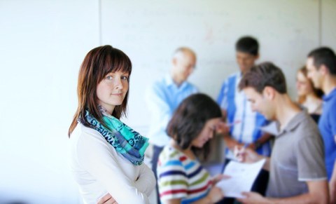 Junge Frau mit dunklen Haaren steht mit verschränkten Armen links im Bild. Im rechten Bildhintergrund befindet sich eine gemischte Gruppe, die sich einander zuwenden.