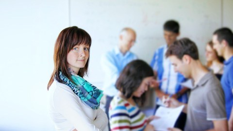 Junge Frau mit dunklen Haaren steht mit verschränkten Armen links im Bild. Im rechten Bildhintergrund befindet sich eine gemischte Gruppe, die sich einander zuwenden.