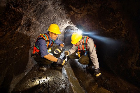 Zwei Männer mit gelben Bauhelmen untersuhchen knieend Wasser in einem Kanal