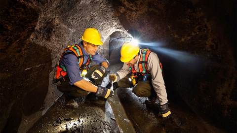 Zwei Männer mit gelben Bauhelmen untersuhchen knieend Wasser in einem Kanal