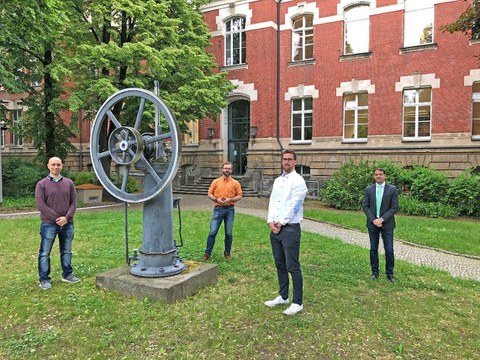 Vier Männer stehen verteilt im Innenhof eines Backsteinbaus, sie umringen ein historisches Modell einer Maschine