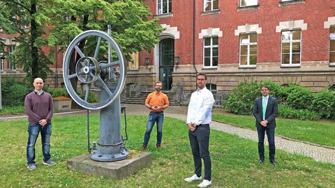 Vier Männer stehen verteilt im Innenhof eines Backsteinbaus, sie umringen ein historisches Modell einer Maschine