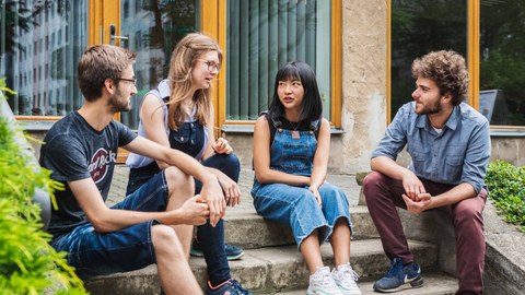 Auf dem Foto sitzen Studenten verschiedener Nationaliäten auf einer Treppe im Freien und unterhalten sich.