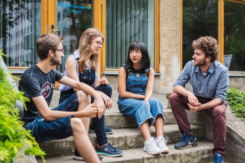 Auf dem Foto sitzen Studenten verschiedener Nationaliäten auf einer Treppe im Freien und unterhalten sich.