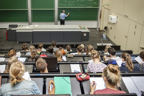 Blick von hinten/oben in eine Vorlesung, vorn steht der Professor und schreibt an eine Tafel.