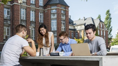 4 Studenten sitzen vor der Biomensa an einem Tisch, vor ihnen steht ein Laptop.