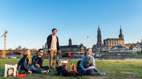 Eine Gruppe junger Menschen sitzt auf der Elbwiese, im Hintergrund die Dresdner Kulisse mit Hofkirche.