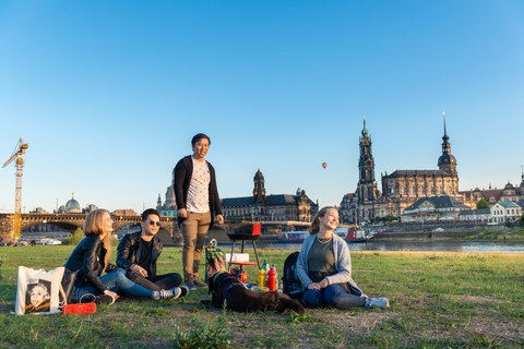 Eine Gruppe junger Menschen sitzt auf der Elbwiese, im Hintergrund die Dresdner Kulisse mit Hofkirche.