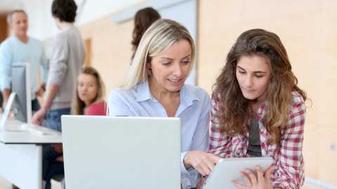 Eine Frau erklärt einer sehr jungen Frau etwas an einem Tablett, vor ihnen steht ein Laptop. Im linken Hintergrund sind vier weitere Personen stehend an einem Pult zu sehen.
