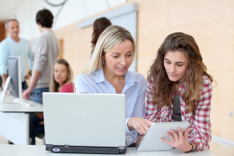 Eine Frau erklärt einer sehr jungen Frau etwas an einem Tablett, vor ihnen steht ein Laptop. Im linken Hintergrund sind vier weitere Personen stehend an einem Pult zu sehen.