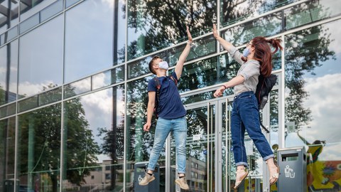 Ein Student und eine Studentin springen vor dem Hörsaalzentrum ein wenig in die Höhe.