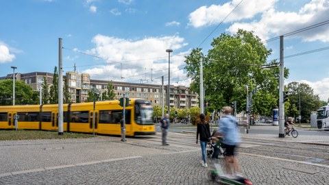 Foto eines großen Platzes, links fährt eine Straßenbahn ins Bild, im rechten Vordergrund sind Passanten zu sehen.