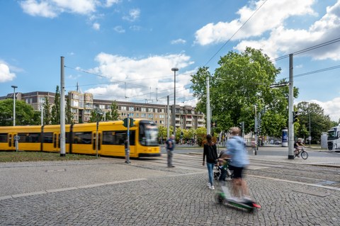 Foto eines großen Platzes, links fährt eine Straßenbahn ins Bild, im rechten Vordergrund sind Passanten zu sehen.