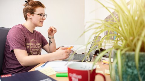 Eine Frau sitzt an einem Schreibtisch, auf dem sich ein Laptop und auch Bücher befinden.