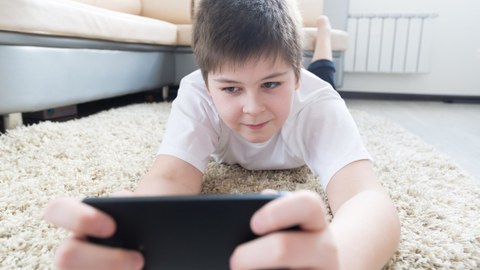 Ein Junge liegt mit dem Kopf nach vorn auf einem hellen Teppich und schaut auf sein Handy