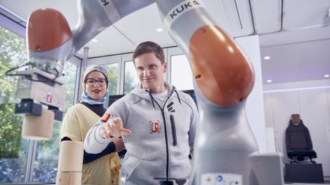 Im Vordergrund ein Roboterarm, der Holzklötzchen anhebt. Im Hintergrund links eine junge Frau mit Brille und Kopftuch schaut zu, rechts ein junger Mann mit kurzen, dunklen Haaren, bedient das Gerät mit über Sensoren, die an seiner Jacke angebracht sind.