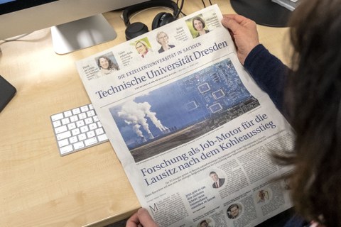 Vor einer hellen Tischplatte hält eine Person eine Zeitungsseite ins Bild.