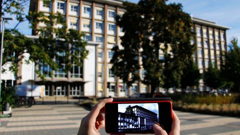 Im Hintergrund der aktuelle Andreas-Schubert-Bau. Im Vordergrund sieht man das historische Gebäude auf einem Smartphone.