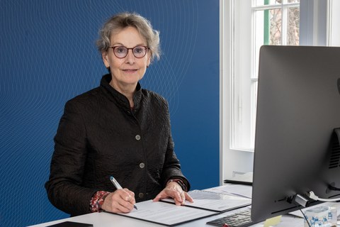 Frau Professorin Staudinger sitzt vor einer blauen Wand, vor sich eine aufgeschlagene Mappe und einen Laptop.