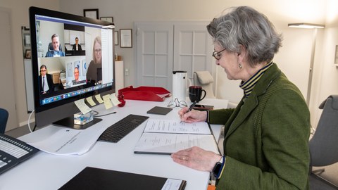 Die Rektorin der TUD sitzt im rechten Bildrand am Schreibtisch und unterzeichnet ein Schriftstück, im Bildschirm gegenüber sind die Teilnehmer der Videokonferenz zu sehen.