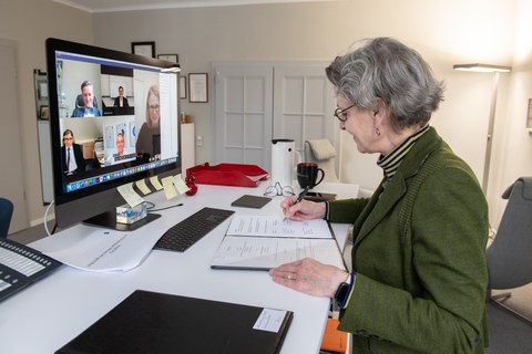 Die Rektorin der TUD sitzt im rechten Bildrand am Schreibtisch und unterzeichnet ein Schriftstück, im Bildschirm gegenüber sind die Teilnehmer der Videokonferenz zu sehen.