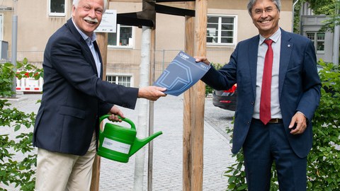 Prof. Müller-Steinhagen steht rechts im Bild und übergibt eine Urkunde an Prof. Bürger, der links im Bild ist. Beide stehen vor dem Baum, den Prof. Bürger gestiftet hat.