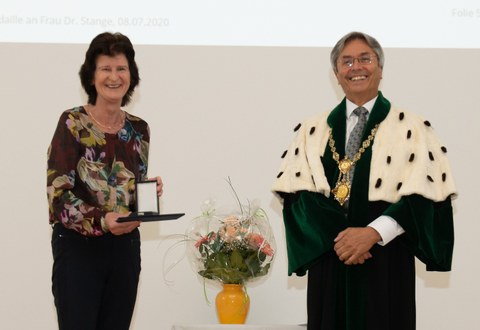 Frau Dr. Stange steht links im Bild, sie hält die Ehrenmedaille in der Hand. Der Rektor steht im Talar rechts. Beide lachen.