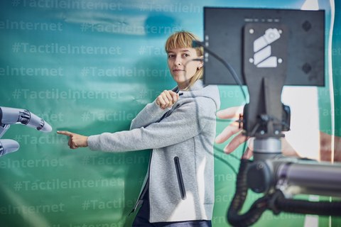 eine junge Frau steht vor einer Wand in blau-grün mit dem mehrfachen Schriftzug #TactileInternet steht und zeigt mit dem Zeigefinger nach vorn.