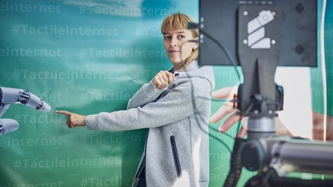 eine junge Frau steht vor einer Wand in blau-grün mit dem mehrfachen Schriftzug #TactileInternet steht und zeigt mit dem Zeigefinger nach vorn.