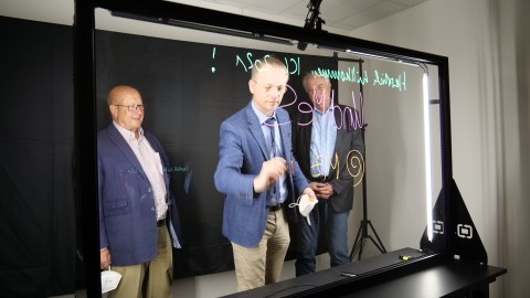 Drei Herren stehen hinter einem durchsichtigen Lightboard, der Herr in der Mitte beschriftet das Board.