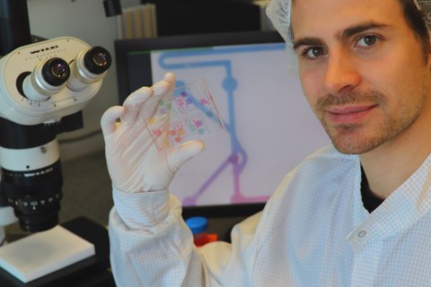 Doktorand Anthony Beck, Mit-Entwickler der Technologie, hält einen chemischen Schaltkreis mit gefärbten Analysemedien. Auf dem Monitor im Hintergrund ist ein Mikroskopiebild eines der chemischen Transistoren des chemischen ICs zu sehen.