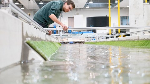 Im Vordergrund ein Wasserkanal in einer Versuchshalle. Im Hintergrund arbeitet ein junger Mann mit Brille an einem Versuchsaufbau im Wasser.
