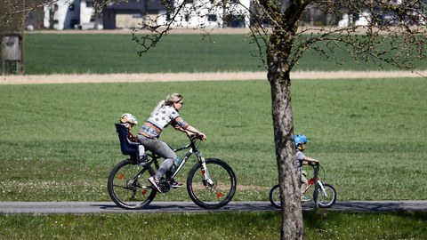 Eine Junge Frau fährt Mountainbike, im Kindersitz hinter ihr sitzt ein Kleinkind, ein weiteres Kind fährt auf seinem Fahrrad vor ihr.