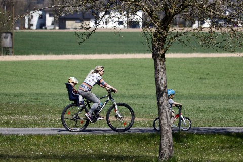 Eine Junge Frau fährt Mountainbike, im Kindersitz hinter ihr sitzt ein Kleinkind, ein weiteres Kind fährt auf seinem Fahrrad vor ihr.