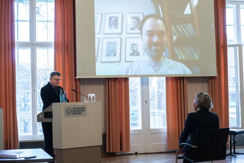 Ein Mann an einem Rednerpult spricht zu einem Mann, der mit Hilfe einer Videokonferenz zugeschaltet ist.