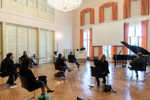 Personen in einem Saal verfolgen die Rede eines Mannes.