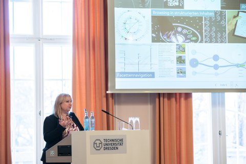 Eine Frau steht an einem Rednerpult und hält einen Vortrag. Im Hintergrund ist eine Prsäentation zu erkennen.