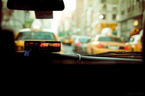 Foto des Innenraums eines Taxis mit Taxometer, man blickt durch die Frontscheibe auf eine start befahrene Straße