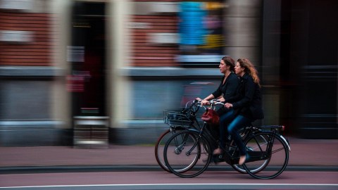 Am rechten unteren Bildrand fahren zwei Frauen auf Fahrrädern nach links, im Hintergrund ist ein Geschäft zu sehen.