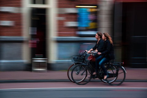 Am rechten unteren Bildrand fahren zwei Frauen auf Fahrrädern nach links, im Hintergrund ist ein Geschäft zu sehen.