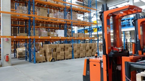 Blick in eine Lagerhalle: Rechts vorn im Bild steht ein orangefarbener Gabelstapler, im Hintergrund stehen deckenhohe, mit Kisten gefüllte Regale  
