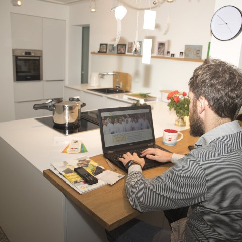 Junger Mann mit Bart sitzt in seiner Küche am Laptop und arbeitet