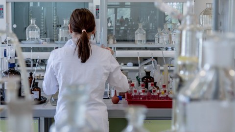Man sieht eine junge Frau mit Zopf von hinten, die inmitten von chemischen Gerätschaften in einem Labor steht.