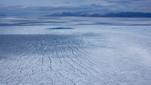 Eine große, rissige Eisfläche, im Hintergrund sind Berge und der Himmel zu sehen, alles in verschiedenen Schattierungen von Hellblau.