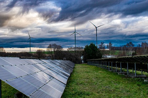 Solarpanele links auf einer Wiese, im Hintergrund sieht man ein Windrad vor bewölktem Himmel.