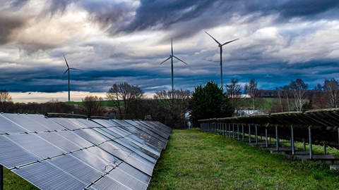 Solarpanele links auf einer Wiese, im Hintergrund sieht man ein Windrad vor bewölktem Himmel.