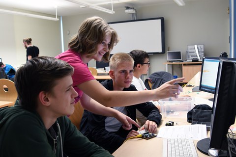 Schüler sitzen nebeneinander an einer Schulbank und sehen auf Bildschirme. Ein Mädchen steht zwischen ihnen und zeigt auf einen Bildschirm.