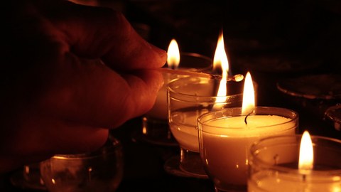 Am rechten Bildrand stehen nebeneinander Teeliche im Dunkeln, von links kommt eine Hand mit STreichholz, die die Kerzen angezündet hat.