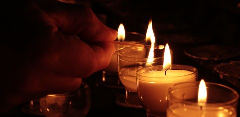 Am rechten Bildrand stehen nebeneinander Teeliche im Dunkeln, von links kommt eine Hand mit STreichholz, die die Kerzen angezündet hat.