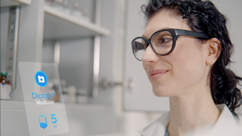 Eine dunkelhaarige junge Frau im rechten Bildrand trägt eine Brille mit dickem schwarzen Rahmen, im Hintergrund ist ein Labor zu sehen.